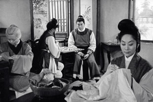 Schwarz-weißes Filmstill zeigt eine stickende Frau im Vordergrund eines Kaffeehauses oder ähnlichem. Im Hintergrund weitere Menschen