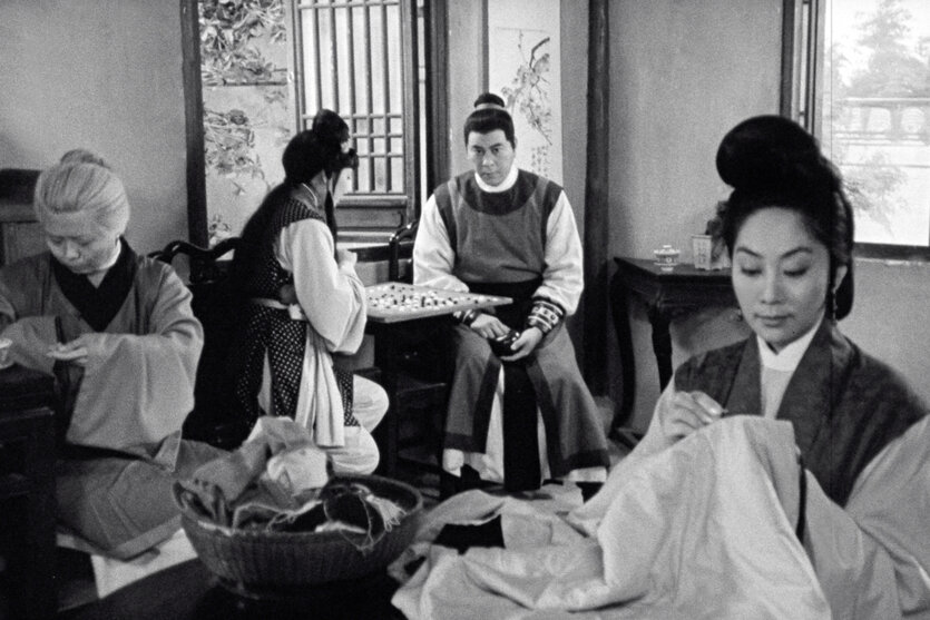 Schwarz-weißes Filmstill zeigt eine stickende Frau im Vordergrund eines Kaffeehauses oder ähnlichem. Im Hintergrund weitere Menschen