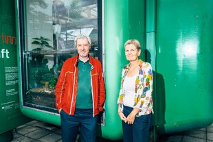 Das Künstlerpaar Adam Page und Eva Hertzsch schauen in die Kamera und lächeln, sie stehen vor einem Space-Age-Gebäude mit knallgrüner Farbe.