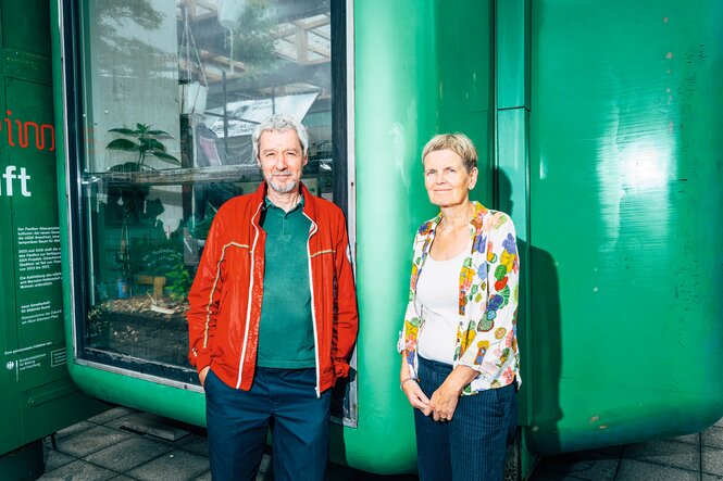 Das Künstlerpaar Adam Page und Eva Hertzsch schauen in die Kamera und lächeln, sie stehen vor einem Space-Age-Gebäude mit knallgrüner Farbe.
