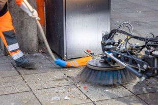 Eine Bürste einer Kehrmaschine rechts im Bild fegt Kippenstummel vom Boden einer Fußgängerzone, links in oranger Montur und mit Besen ein städtischer Mitarbeiter.