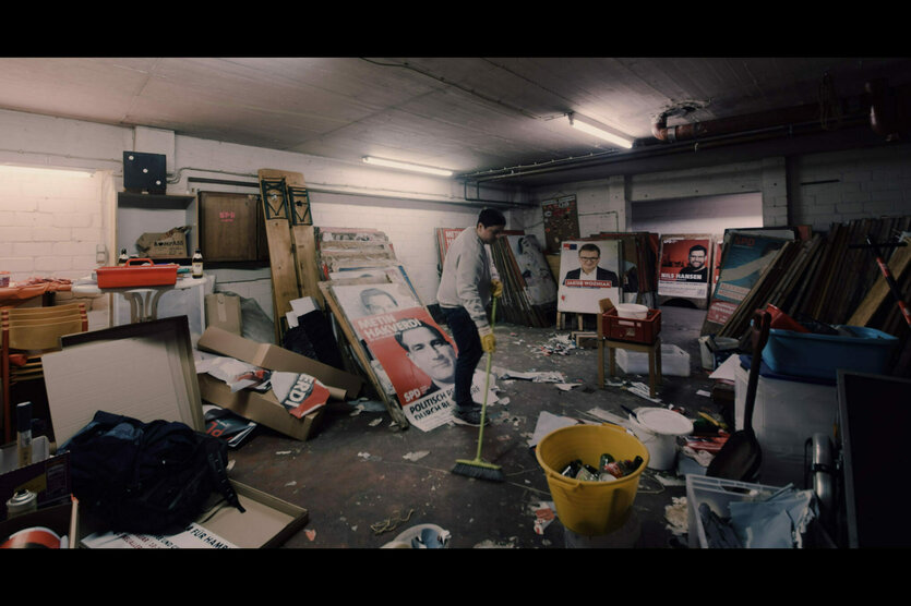 Filmszene: Ein Mensch fegt einen Raum voller Wahlplakate