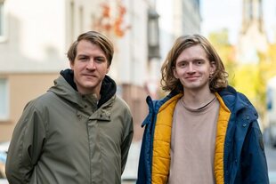 Zwei Männer, beide 28 Jahre alt und blond, draußen auf der Straße. Links Artem Klyga, trägt einen Scheitel und eine dunkelbeige Jacke. Rechts Sascha Belik, gewellte Haare bis kurz über die Schulter, seine geöffnete Jacke ist außen Blau, innen gelb.