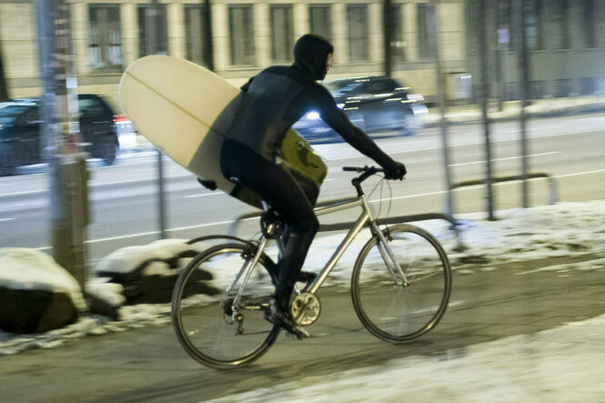 Ein Mann im Neoprenanzug radelt bei Schnee mit seinem Surfboard untern dem Arm