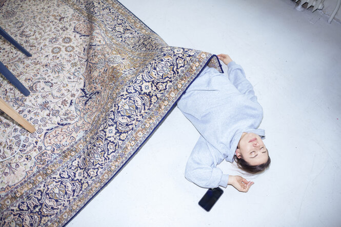 eine Frau liegt auf dem Boden und ist mit einem Teppich zugedeckt, neben ihr liegt ein Handy