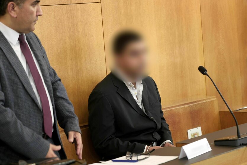 Ein Mann sitzt in einem Gerichtssaal, neben ihm steht ein Mann in Anzug