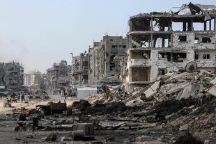 Auf dem Bild ist eine stark zerstörte Straße mit eingestürzten Häusern und Trümmern zu sehen. Einige Menschen gehen zwischen den Ruinen entlang.