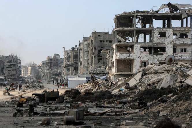 Auf dem Bild ist eine stark zerstörte Straße mit eingestürzten Häusern und Trümmern zu sehen. Einige Menschen gehen zwischen den Ruinen entlang.