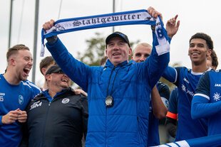 Mann in blauer Jacke hält blauen Schal hoch, auf dem Schal steht in Fraktur 