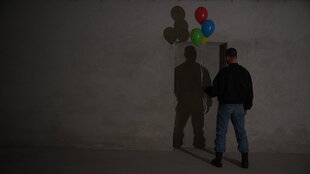 Ein Mann steht mit bunten Ballons vor einer grauen Wand