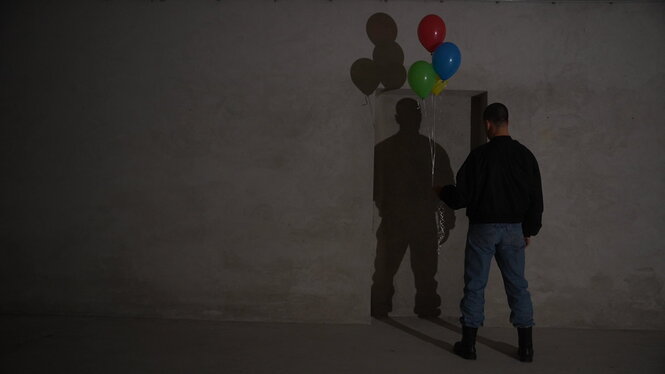 Ein Mann steht mit bunten Ballons vor einer grauen Wand