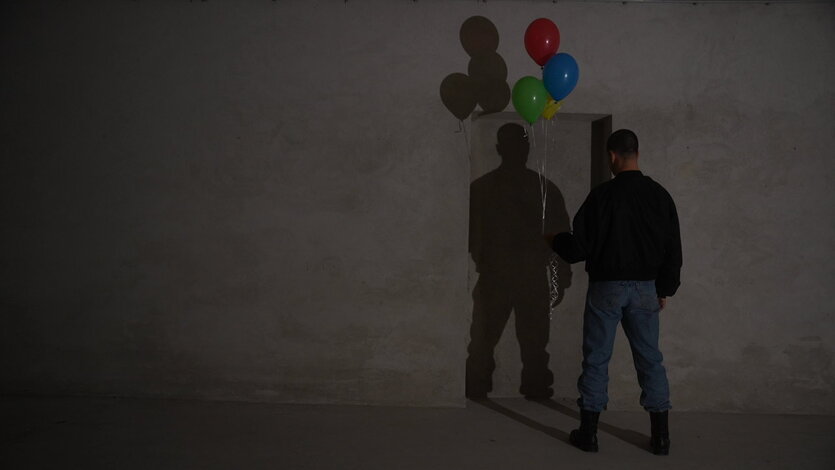 Ein Mann steht mit bunten Ballons vor einer grauen Wand