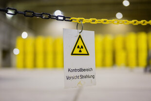 In einer Lagerhalle stehen aufeinandergestapelt zwei Reihen gelbe Metallfässer. Davor hängt an einer gelb-schwarzen Absperrkette ein Warnschild mit dem Symbol der Atomkraft und den Worten: "Kontrollbereich Vorsicht Strahlung".