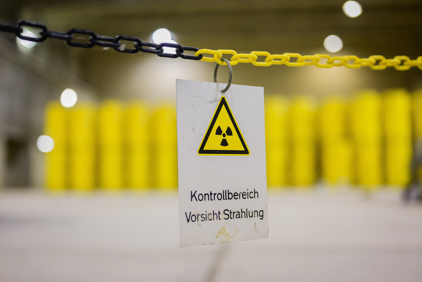 In einer Lagerhalle stehen aufeinandergestapelt zwei Reihen gelbe Metallfässer. Davor hängt an einer gelb-schwarzen Absperrkette ein Warnschild mit dem Symbol der Atomkraft und den Worten: "Kontrollbereich Vorsicht Strahlung".