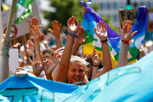 Mehrere Kinder laufen in einer Reihe draußen zwischen wehenden bunten Fahnen und Bannern. Sie recken ihre Arme nach oben. Im Fokus der Kamera ist ein lachendes Kind mit Pferdeschwanz.