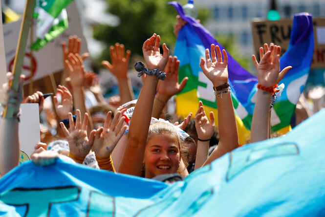 Mehrere Kinder laufen in einer Reihe draußen zwischen wehenden bunten Fahnen und Bannern. Sie recken ihre Arme nach oben. Im Fokus der Kamera ist ein lachendes Kind mit Pferdeschwanz.