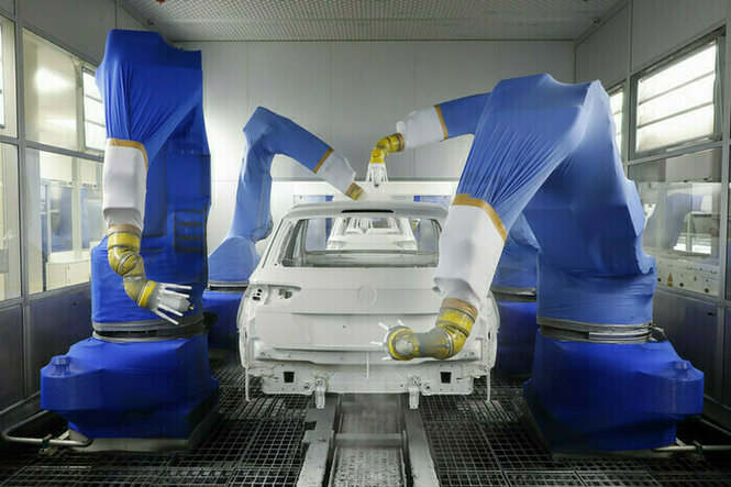 Der Rohbau eines VW-Autos wird maschinell lackiert. Die Maschine sieht aus, als habe sie lange Arme, überzogen mit einem blauen Schutzanzug. Das Auto ist bisher weiß lackiert.