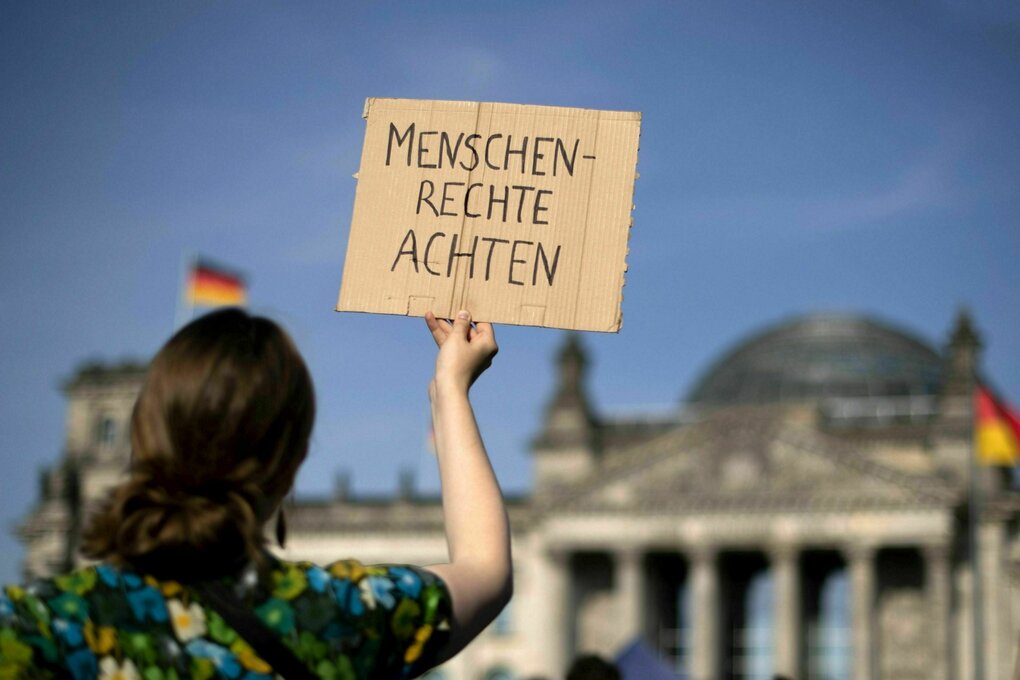 Eine Person hät ein Protestschild hoch, auf dem steht: MENSCHENRECHTE ACHTEN, im Hintergrund das Reichstagsgebäude