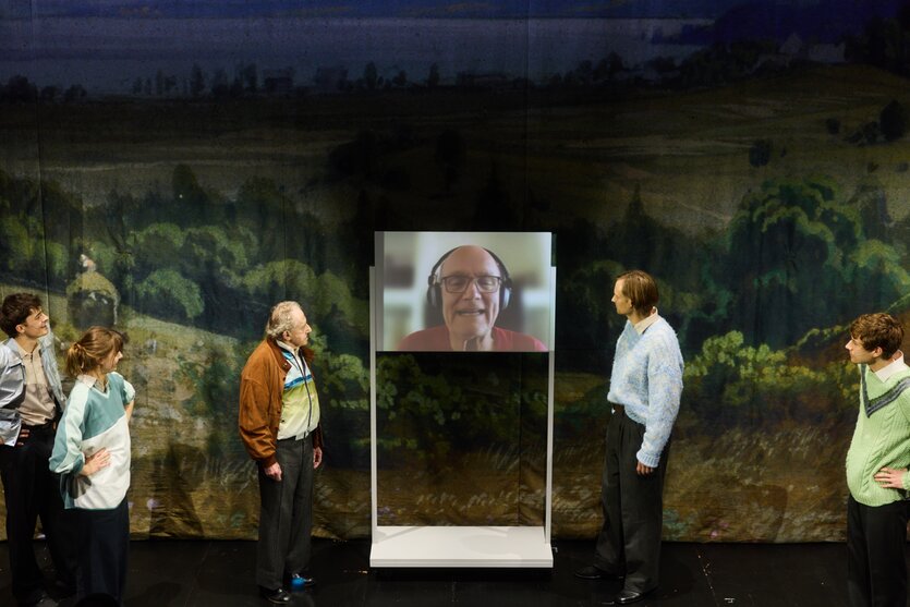 Blick auf die Bühne des Stücks „Zeit ohne Gefühle“ mit mehreren Schauspielern, die um einen Bildschirm stehen, auf dem ein Mann höheren Alters per Videoschalte am Geschehen teilnimmt