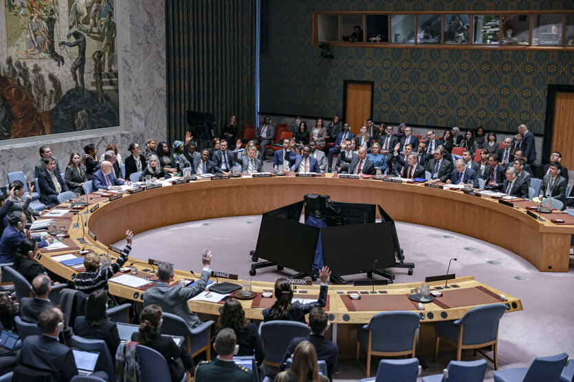 In einem großen Konferenzsaal sitzen viele Menschen um einen runden Tisch herum. Einige heben ihre Hände