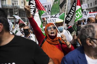 Eine Menschengruppe läuft in einem Demonstrationszug zwischen Gebäuden hindurch. Einige tragen Fahnen in der der Hand. In der Mitte läuft eine Frau, die eine Faust nach oben streckt und etwas ruft