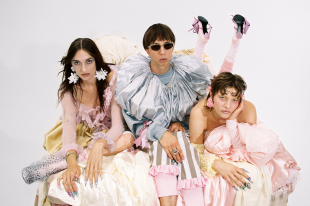 Das Bild zeigt die drei Bandmitglieder von Blond in extravaganten, silbernen und pastellfarbenen Outfits, die auf einem Haufen Stoffe vor weißem Hintergrund sitzen und liegen.