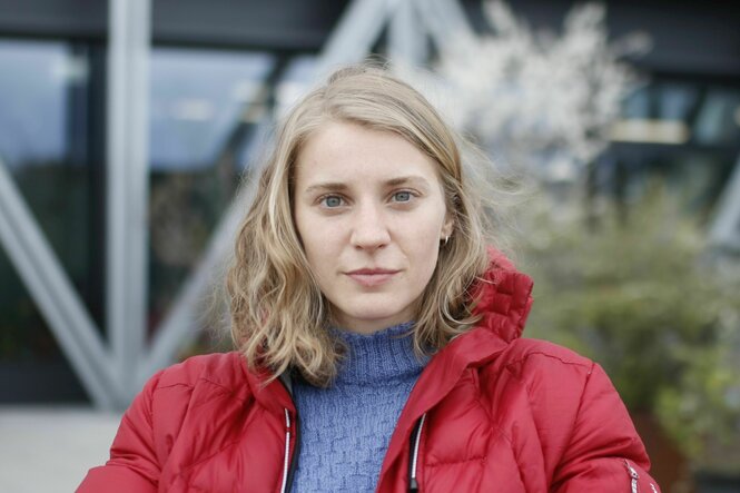 Porträt von Luisa Faust mit roter Jacke, einem lilanen Rollkragenpullover und schulterlangen, blonden Haaren, der Blick ist direkt in die Kamera. Im Hintergrund sind ein modernes Gebäude mit Stahlstreben.