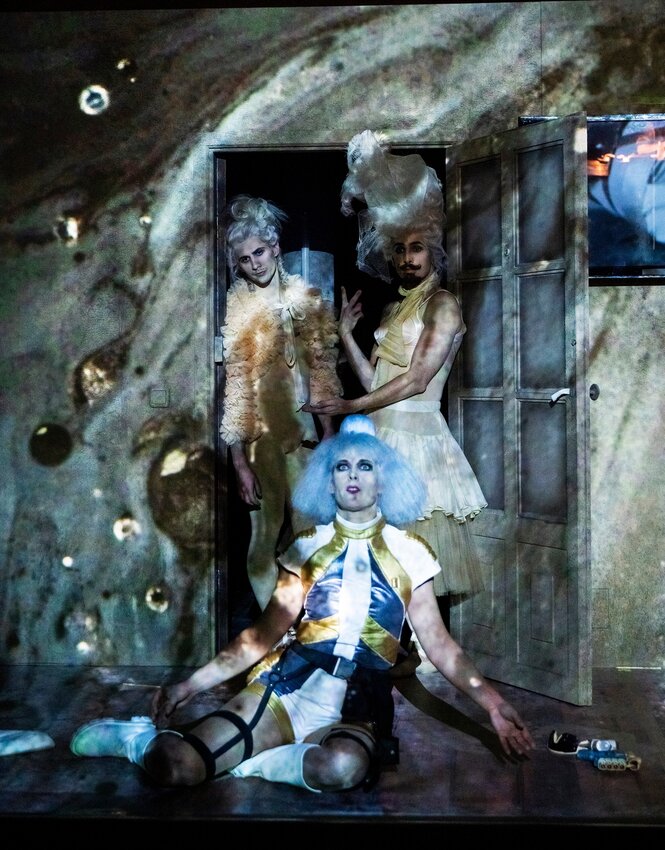 Zwei Figuren mit hohen Perücken stehen in einer Tür, davor sitzt eine auf dem Boden