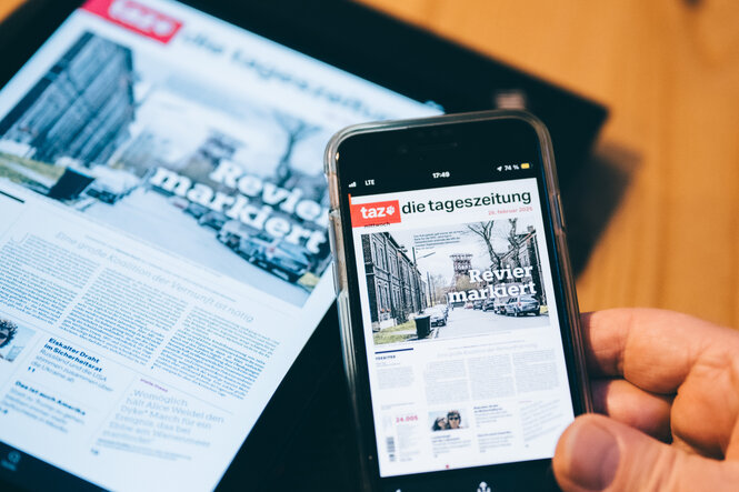 Eine Person hält ein Smartphone in der Hand, auf dem die Titelseite der digitalen Ausgabe der taz angezeigt wird. Im Hintergrund ist dieselbe Titelseite auf einem Tablet zu sehen. Die Schlagzeile lautet „Revier markiert“ und zeigt ein Foto einer Straße mit geparkten Autos und alten Backsteingebäuden.