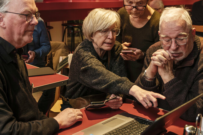 Vier ältere Personen sitzen eng beieinander an einem Tisch in der taz Kantine und schauen konzentriert auf einen Laptop. Eine Frau in der Mitte zeigt mit dem Finger auf den Bildschirm, während die anderen aufmerksam zuhören und mitdenken. Smartphones lieg