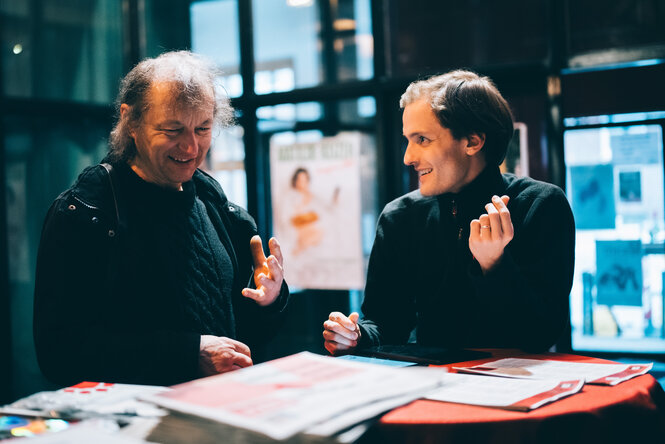 taz-Seitenwende-Tourmanager Vincent Bruckmann-Levinson (re.) und ein Leser stehen an einem kleinen Tisch in einem Foyer und unterhalten sich angeregt. Beide lächeln, eine Person gestikuliert mit den Händen, die andere hält ein Papier in der Hand. Auf dem Tisch liegt die taz, ein Tablet und Flyer. Die Atmosphäre wirkt freundlich und lebhaft.
