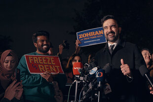 Zu sehen ist der New Yorker Bürgermeisterkandidat Zohran Mamdani. Hinter ihm hält jemand ein Schild mit der Aufschrift "Zohran for New York City" in die Luft, daneben sieht man einen lächelnden Mann, mit einem aktivistisch anmutenden Schild, auf dem "Freeze the rent" zu lesen ist.