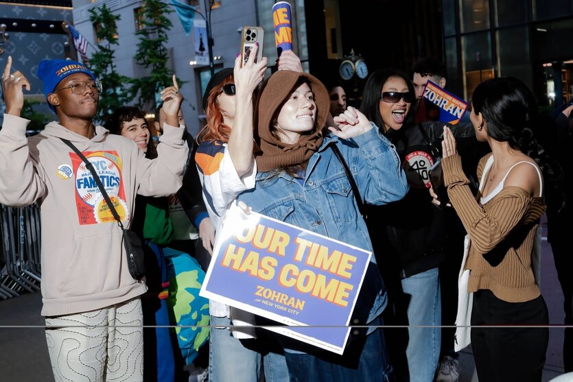 Junge Anhänger und Anhängerinnen von Zohran Mamdani stehen am Straßenrand und jubeln, eine trägt ein Plakat mit der Aufschrift "Our time has come"