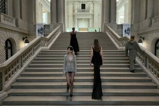 Mehrere Models schreiten auf einer breiten, monumentalen Steintreppe in einem prunkvollen historischen Gebäude hinauf und hinab. Sie tragen minimalistische anmutende Mode in gedeckten Farbtönen..