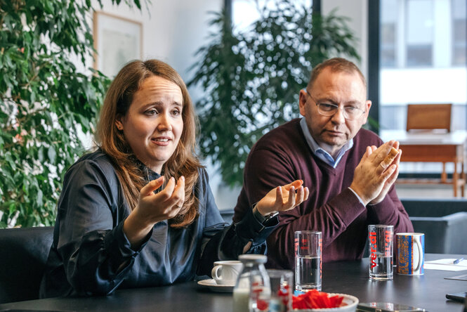 Ricarda Lang sitzt mit Frank Werneke am Tisch und diskutiert, sie hält die Hände fragend vor sich - hinter ihnen steht ein Ficus Benjamini