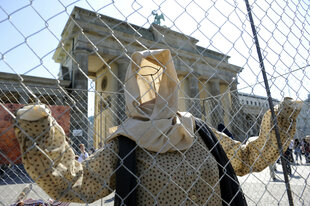 Die Kleidung einer Person mit Kopftuch ist vor dem Brandenburger Tor an einem Zaun befestigt, dadurch ergibt sich die Silhouette eines Menschen mit am Gitter hochgehaltenen Händen.