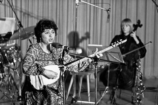 Schwarzweißbild einer Frau mit einer Langhalslaute auf einer Bühne, vor ihr ein Mikrofonständer