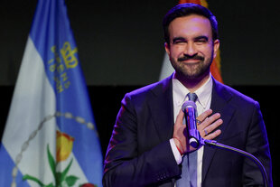 Ein Mann im Anzug hält sich die Hand vor die Brust und grinst