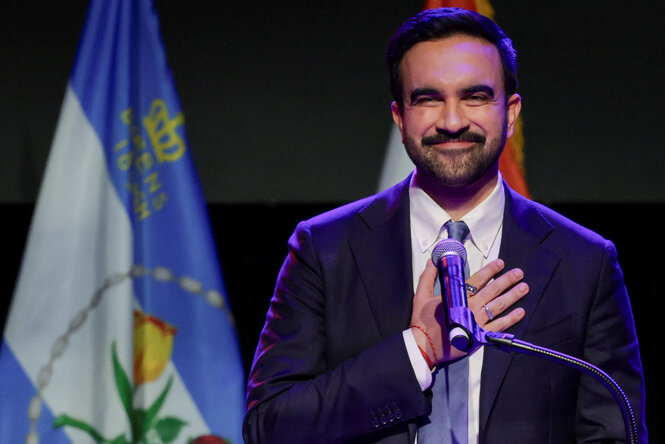 Ein Mann im Anzug hält sich die Hand vor die Brust und grinst