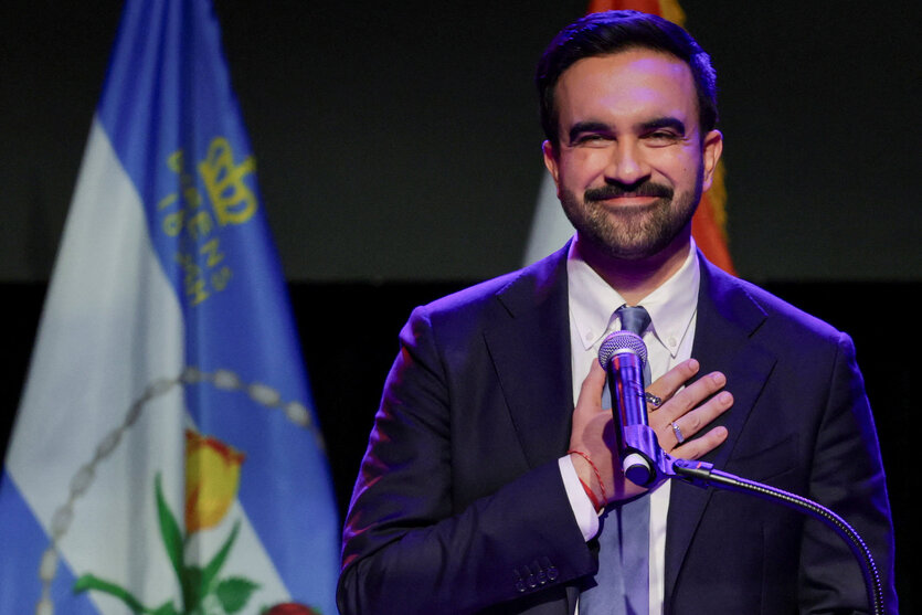 Ein Mann im Anzug hält sich die Hand vor die Brust und grinst
