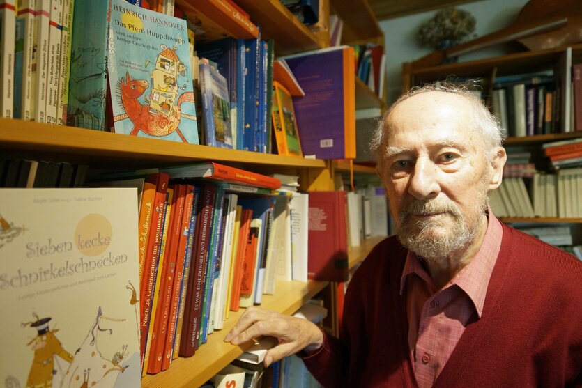 Auf dem Bild ist ein älterer Mann mit grauen Haaren und Bart zu sehen. Er trägt ein rotes Hemd und eine rote Strickjacke. Er steht in einem Raum voller Bücherregale, die dicht mit bunten Büchern gefüllt sind. Einige Bücher sind Kinderbücher mit farbenfrohen Covern und Illustrationen. Der Mann schaut ruhig und freundlich in die Kamera.