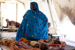 Eine Frau sitzt in einem Zelt, sie schützt sich mit einer blauen Decke, auf dem Boden vor ihr liegen kleine Kinder auf Matrazen