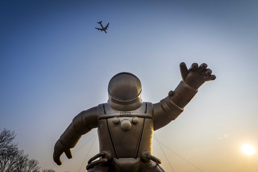 Skulptur eines Astronautenanzugs vor Abendhimmel, ein Flugzeug fliegt vorbei