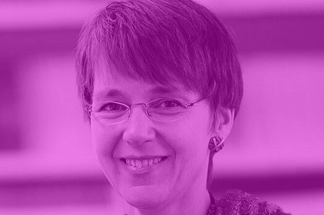 Portät von Nicole Mayer-Ahuja mit randloser Brille, einem Holzohrring, leicht lächelnd und kurzen graumelierten Haaren.