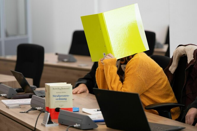 Eine Frau, die einen gelben Pullover trägt, hält sich im Gerichtssaal einen gelben Aktenordner vors Gesicht