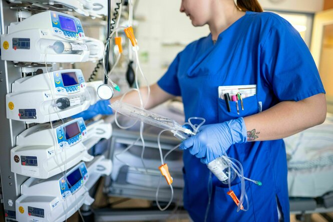 Eine junge Frau steht in blauer Arbeitskleidung vor Apparaten, im Hintergrund ein Krankenbett