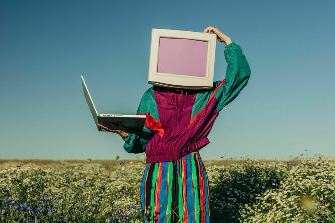 Eine Person mit einem Monitor als Kopf und einem Laptop im Arm steht auf einer Wiese