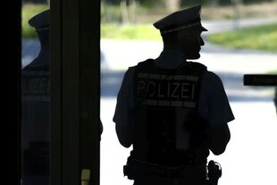 Zwei Silhouetten von hinten: Beamte mit Mütze und Weste mit Polizei-Aufschrift auf dem Rücken. Der Blick geht durch ein Glasfenster, die Gesichter sind nicht zu erkennen.