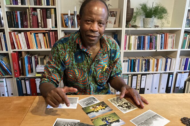 Ein Mann sitzt hinter einem Schreibtisch vor einem Bücherregal, vor sich Fotos ausgebreitet