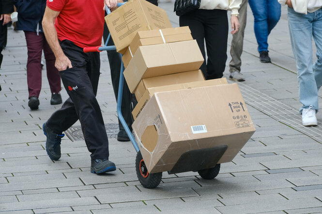 Ein Paketbote transportiert gestapelte Pakete auf einer Sachkarre auf einem Gehweg